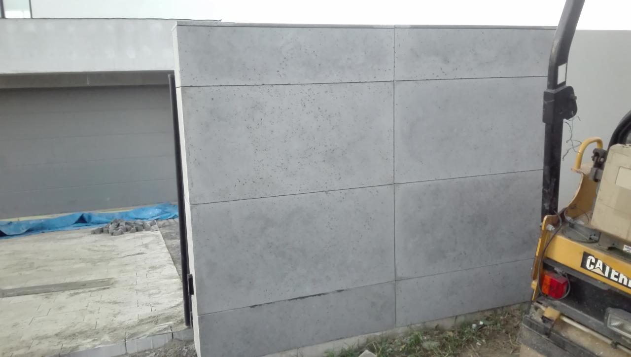Szara ściana z prefabrykatów betonowych w trakcie budowy, widoczna zagęszczarka w tle i przygotowane kostki brukowe.