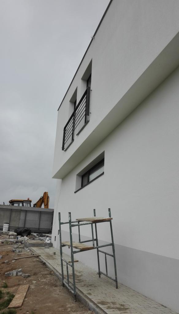 Fragment elewacji budynku z pomalowaną na biało górną częścią i szarą dolną, widoczne rusztowanie, okna z czarnymi ramami, balkony z czarną balustradą oraz koparka w tle.
