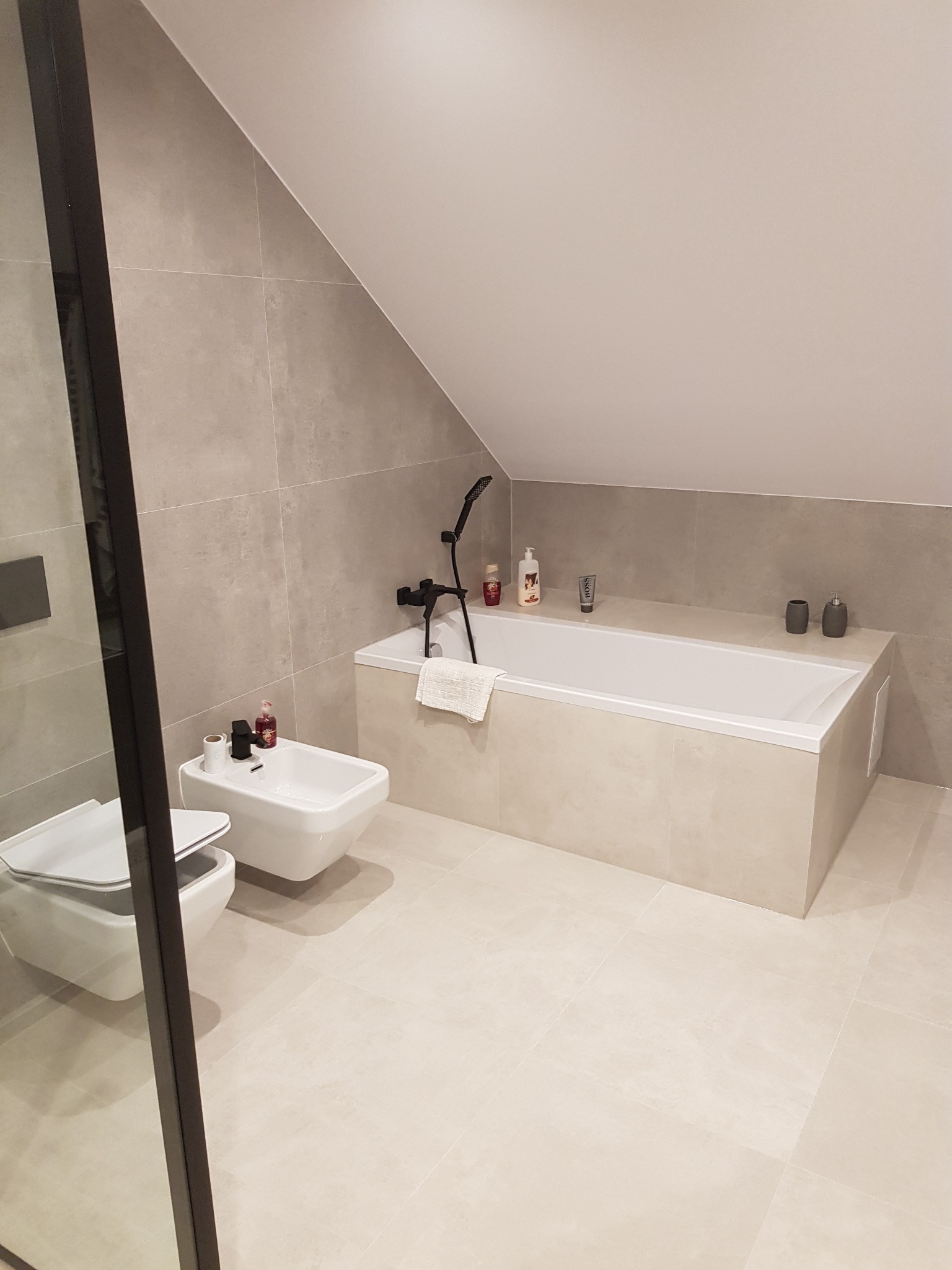 Wykończona łazienka na poddaszu z beżowymi płytkami, białą wanną obudowaną płytkami, bidetem i toaletą. Czarna armatura i szklana kabina prysznicowa.