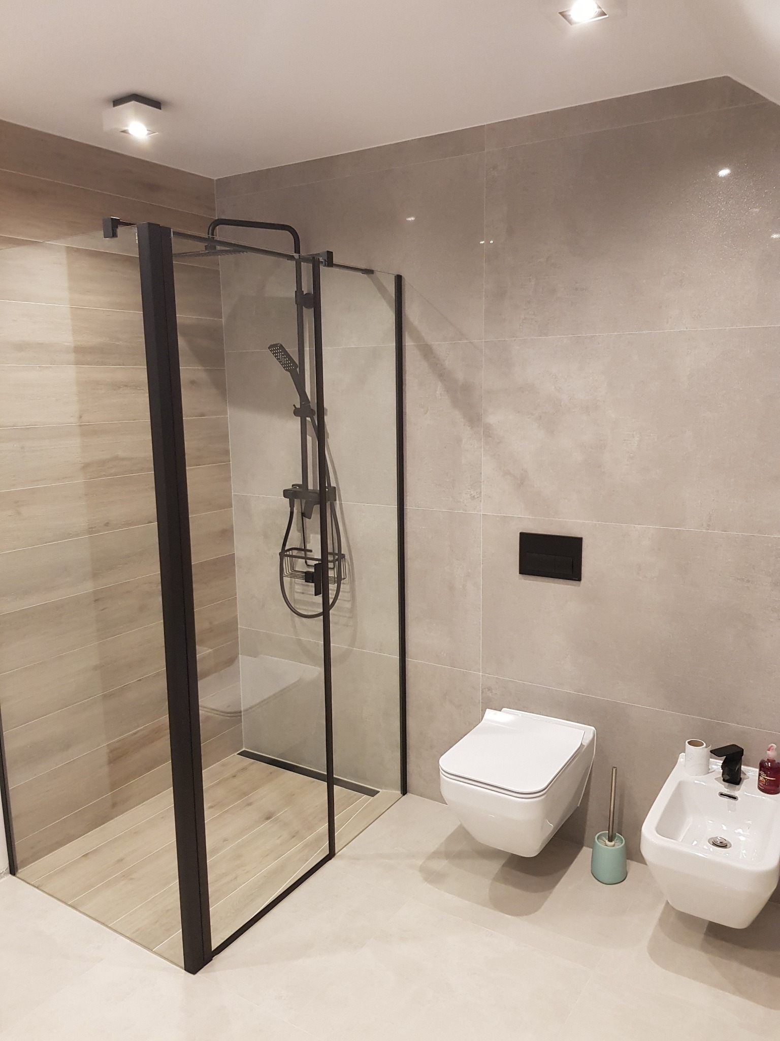 Nowoczesna łazienka z kabiną prysznicową typu walk-in z czarnymi profilami, wiszącą toaletą i bidetem, wykończona płytkami w odcieniach szarości i beżu, oświetlona punktowymi lampami sufitowymi.