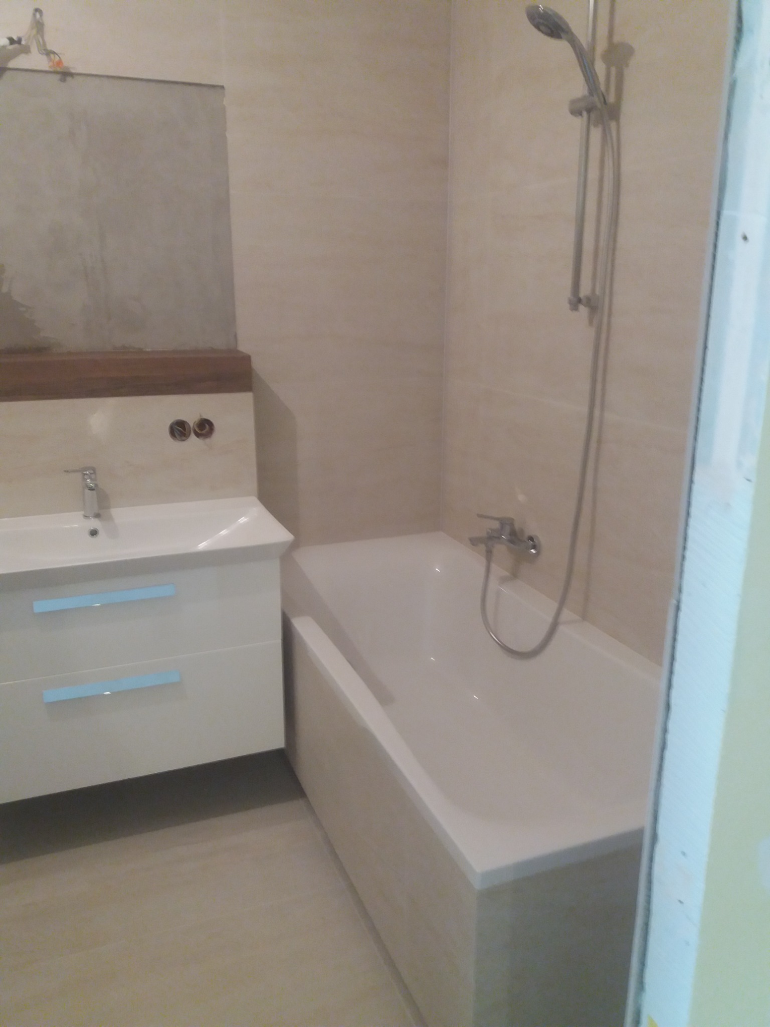 Wykończona łazienka z beżowymi płytkami, białą wanną i umywalką z szafką; widoczne niezabezpieczone kable elektryczne nad umywalką.