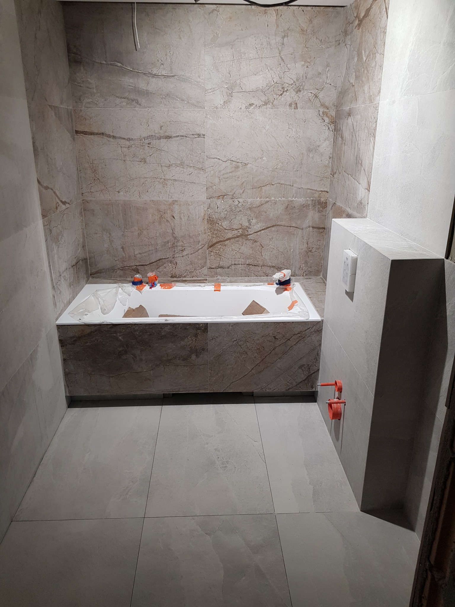 Łazienka w trakcie wykańczania: ściany i obudowa wanny wyłożone płytkami imitującymi marmur, na wannie zabezpieczenia, widoczne przyłącza wodne i częściowo ułożona podłoga z szarych płytek.