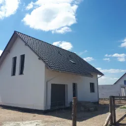 Nowoczesny dom jednorodzinny z grafitową dachówką ceramiczną, widoczne orynnowanie i okno dachowe, w tle błękitne niebo z chmurami, teren wokół domu w trakcie prac wykończeniowych.