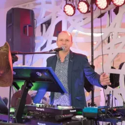 Mężczyzna w marynarce i wzorzystej koszuli śpiewa do mikrofonu, obsługując konsoletę Numark, z głośnikiem w tle i kapeluszem kowbojskim zawieszonym po lewej stronie.