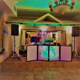 Sala bankietowa z rozstawionym sprzętem DJ-skim: konsoleta z oświetleniem, dwa głośniki na statywach, lustro w ozdobnej ramie w tle, parkiet z płytek.