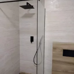 Nowoczesna kabina prysznicowa typu walk-in z czarną armaturą, imitująca drewno płytka na podłodze i ścianie, widoczny odpływ liniowy