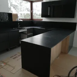 Czarna, matowa zabudowa kuchenna w kształcie litery U z blatem, widok na okno z zimowym krajobrazem, podłoga zabezpieczona kartonem, widoczne elementy instalacji elektrycznej i rura odkurzacza.