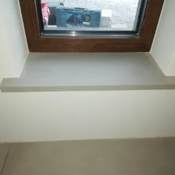 Zamontowany parapet z jasnego konglomeratu w oknie z brązową ramą, widoczna skrzynka narzędziowa za oknem.