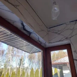 Wykończenie okna wewnątrz marmurem z czarnymi żyłami, widok na zewnątrz przez okno, lampa w folii ochronnej na suficie.