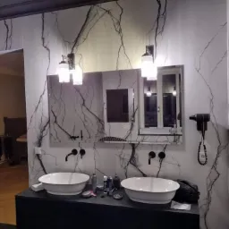 Elegancka łazienka z umywalkami nablatowymi, czarną armaturą i ścianami wykończonymi płytami imitującymi marmur z wyraźnym, czarnym żyłowaniem, dwie lampy ścienne z białymi kloszami i suszarka...