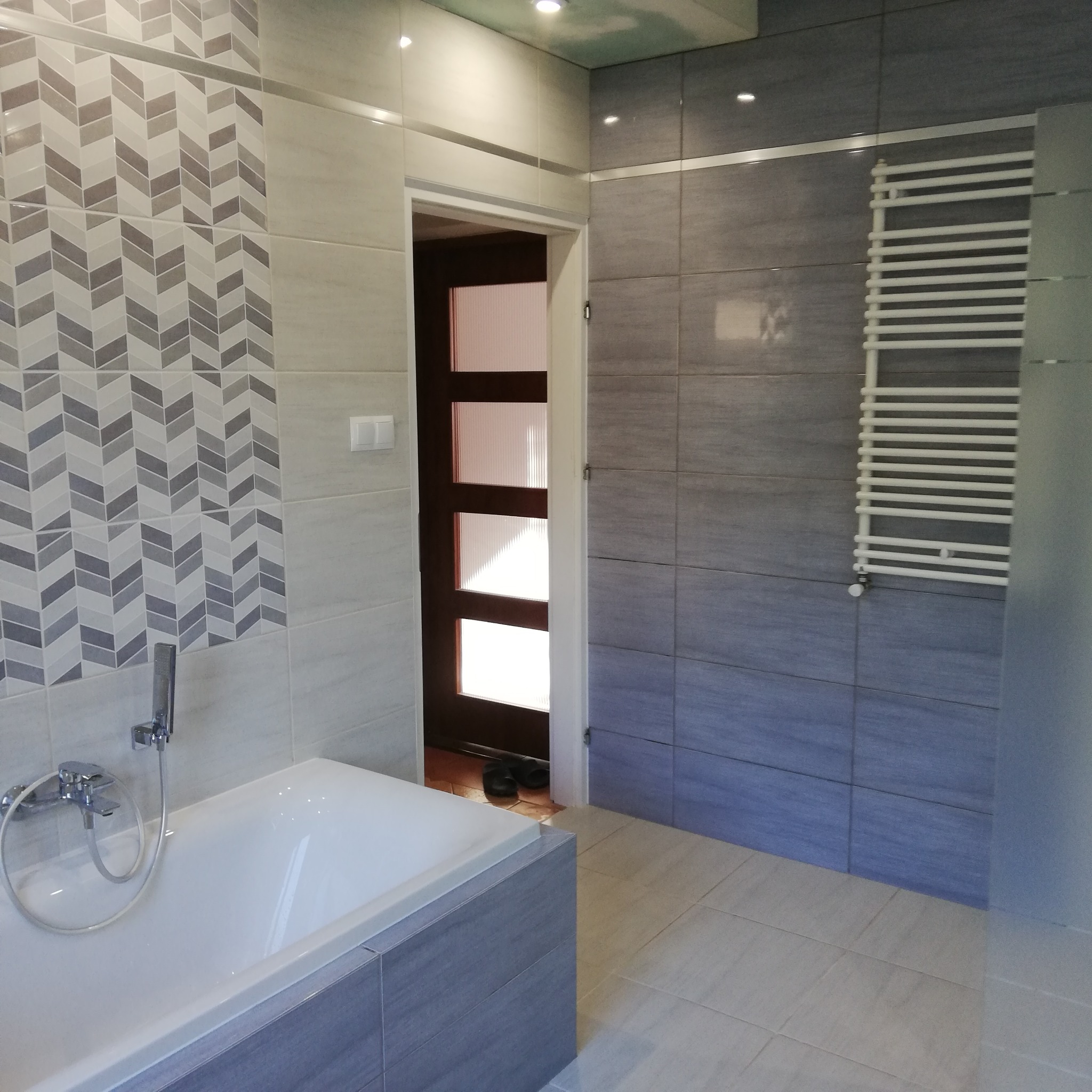 Nowoczesna łazienka w odcieniach szarości i bieli z geometrycznym wzorem płytek nad wanną, grzejnik drabinkowy na ścianie i widoczne kapcie w progu drzwi.