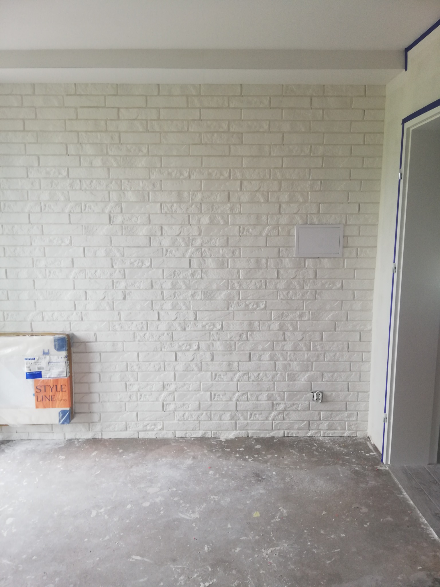 Ściana wykończona białą cegłą dekoracyjną z widoczną puszką elektryczną i zabezpieczoną folią podłogą, w trakcie prac wykończeniowych.