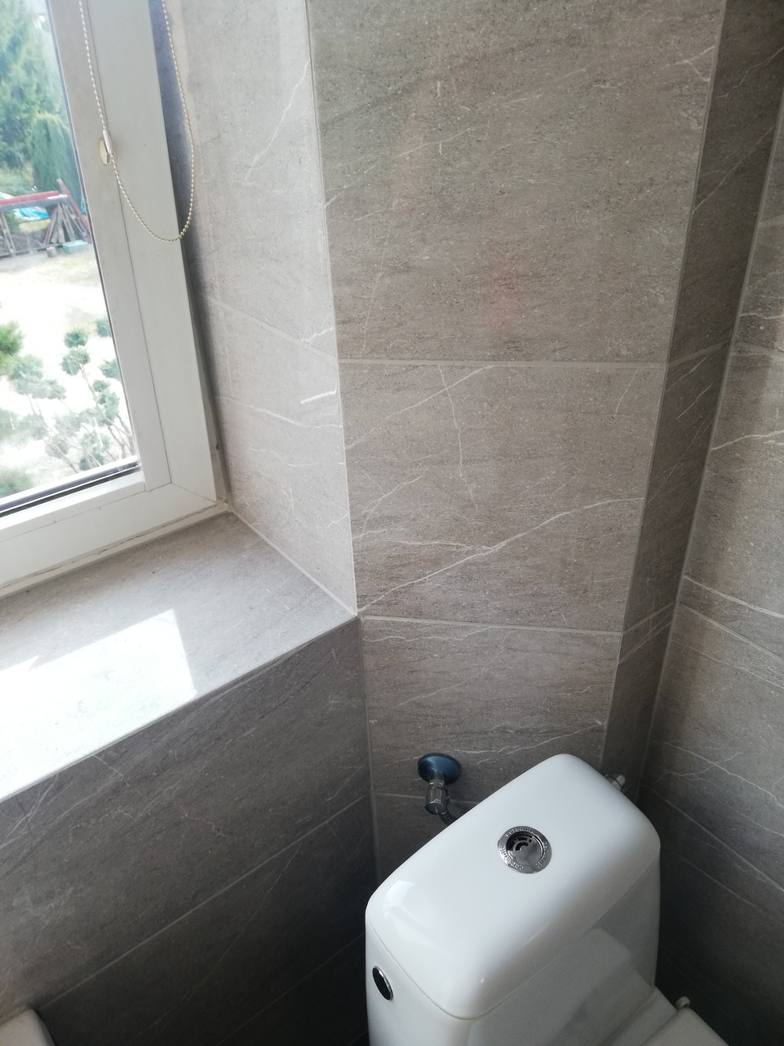 Szare płytki imitujące marmur na ścianie łazienki, widoczny fragment okna z roletą i biała toaleta
