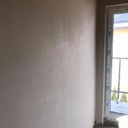 Świeżo nałożony, lekko połyskujący gładki tynk na ścianie wewnętrznej, widoczne ślady po szpachlowaniu, okno PCV w tle.