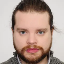 Portret mężczyzny z brodą i włosami upiętymi w kok, ubrany w szary sweter i koszulę w paski, na jasnym tle. Ujęcie centralne, neutralny wyraz twarzy.