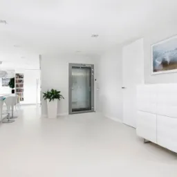 Minimalistyczne wnętrze z białą, błyszczącą podłogą, windą osobową w głębi i nowoczesną komodą z obrazem na ścianie, oświetlone punktowymi lampami.