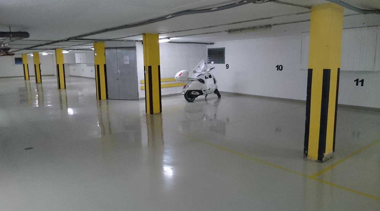 Jasnoszara, błyszcząca posadzka w garażu podziemnym z żółto-czarnymi słupami i zaparkowanym białym skuterem.