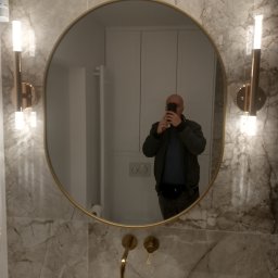 Andrzej Kołomyjski Wykończenia Wnętrz - Owalne lustro w złotej ramie, odbijające fragment łazienki z marmurowymi płytkami, złotą armaturą i białym umywalką, fotografowane przez osobę z telefonem.