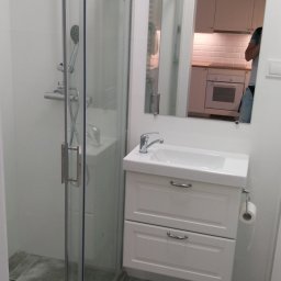 Andrzej Kołomyjski Wykończenia Wnętrz - Biała łazienka z kabiną prysznicową, umywalką z szafką i lustrem, w którym odbija się fragment kuchni.