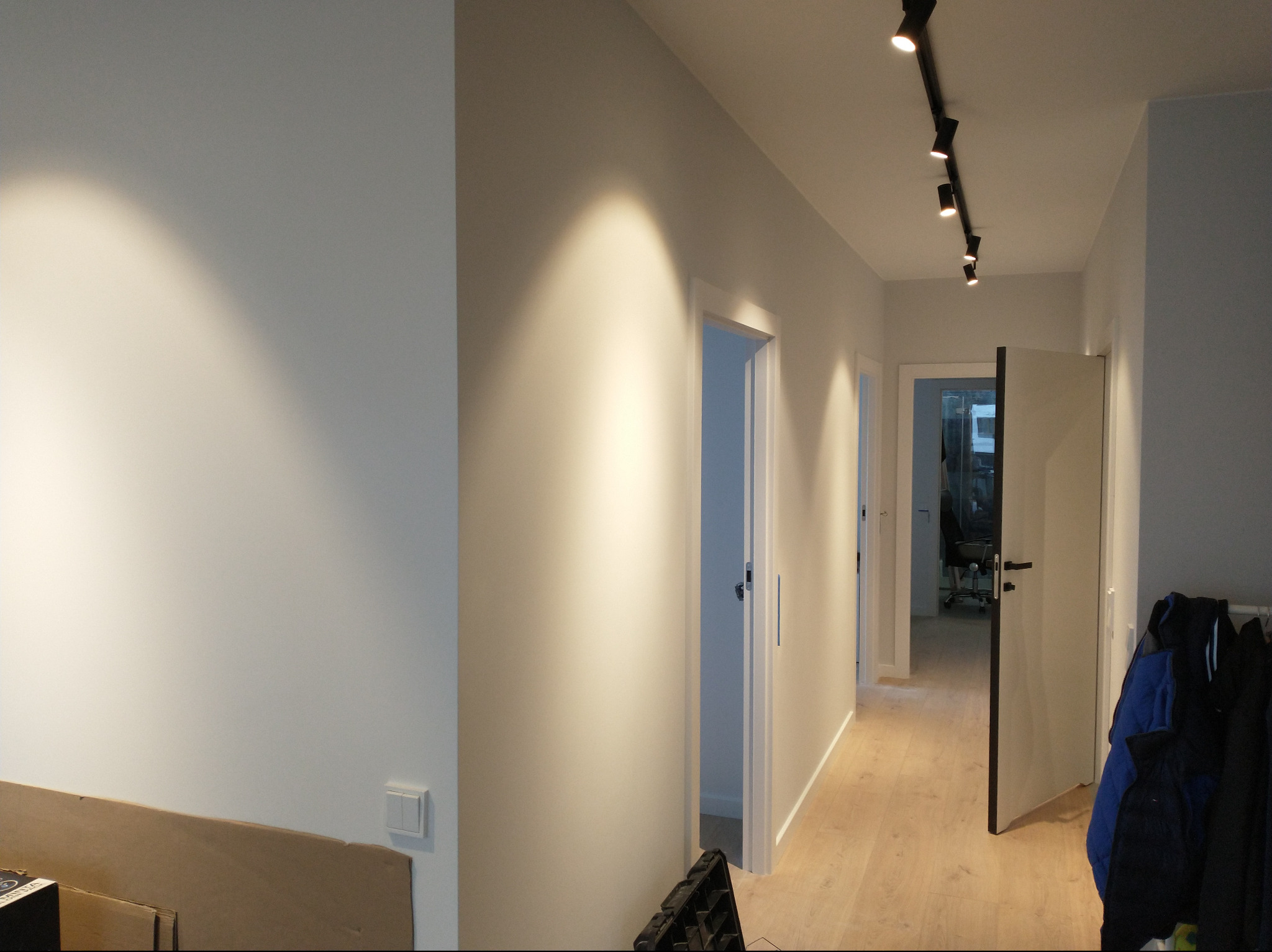 Korytarz po remoncie z białymi ścianami, drewnianą podłogą i oświetleniem szynowym z reflektorami punktowymi.