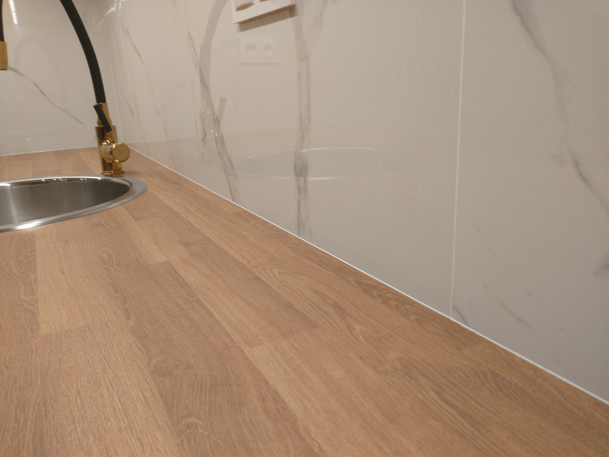 Fragment wykończonej kuchni: drewniany blat, stalowy zlew z czarną baterią z elementami złota, ściana wyłożona płytkami imitującymi marmur z widocznymi gniazdkami elektrycznymi.