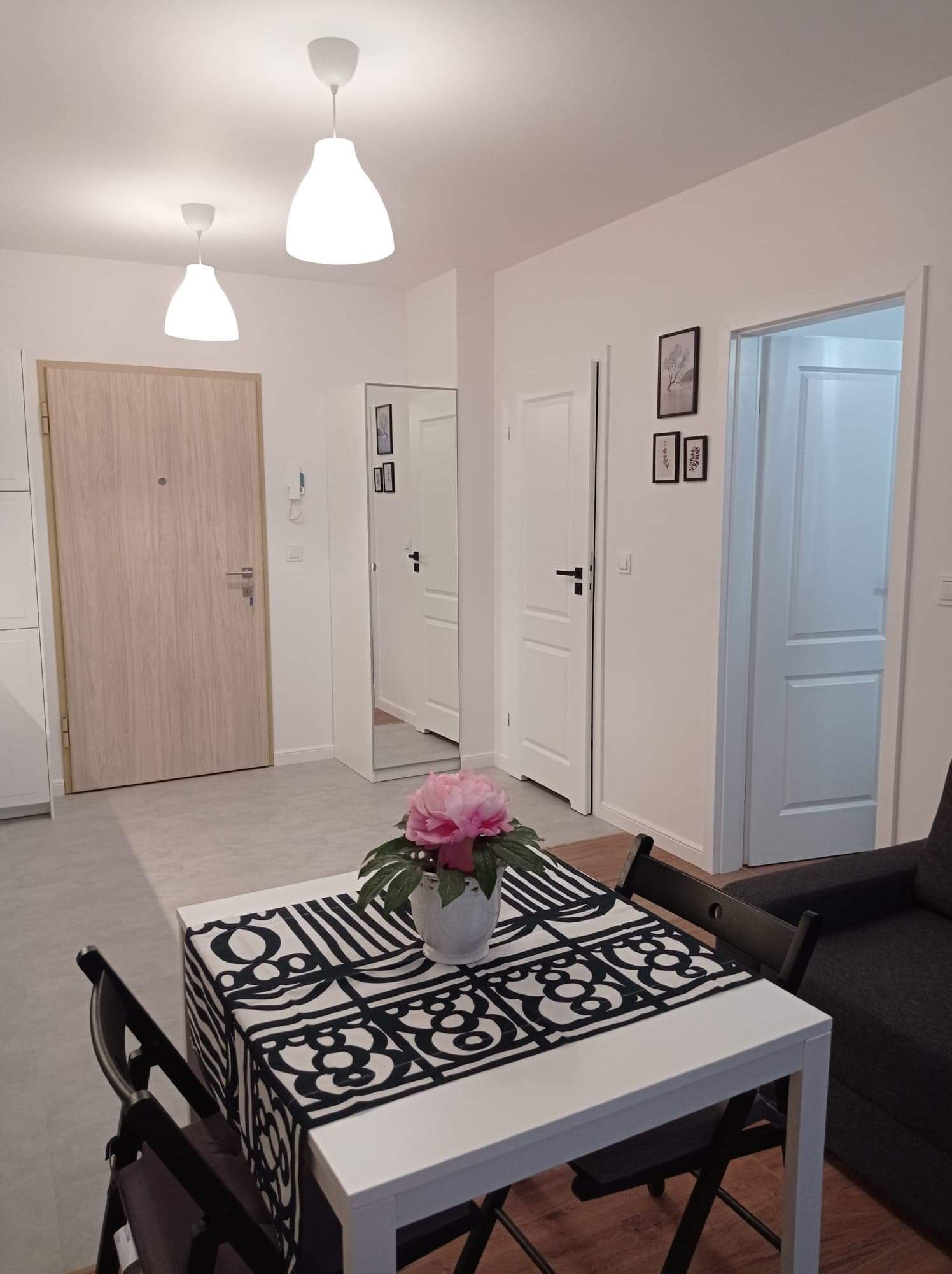 Jasne wnętrze mieszkania po remoncie: białe ściany, drzwi, podłoga w odcieniach szarości, stół z czarno-białym bieżnikiem i bukietem różowych kwiatów w wazonie, widoczne lampy sufitowe.