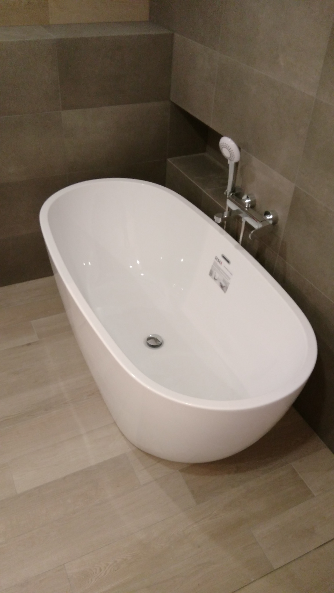 Wolnostojąca, biała wanna w łazience wykończonej płytkami w odcieniach brązu i beżu, z baterią i słuchawką prysznicową. Realizacja w Warszawie.