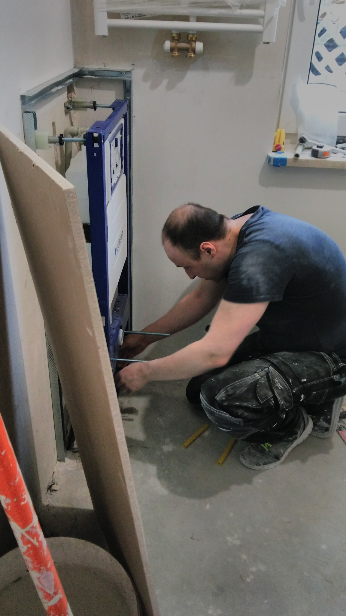 Instalator przykręca stelaż podtynkowy WC Geberit Duofix w łazience podczas remontu, widoczne rury instalacyjne, płyta gipsowa i narzędzia.