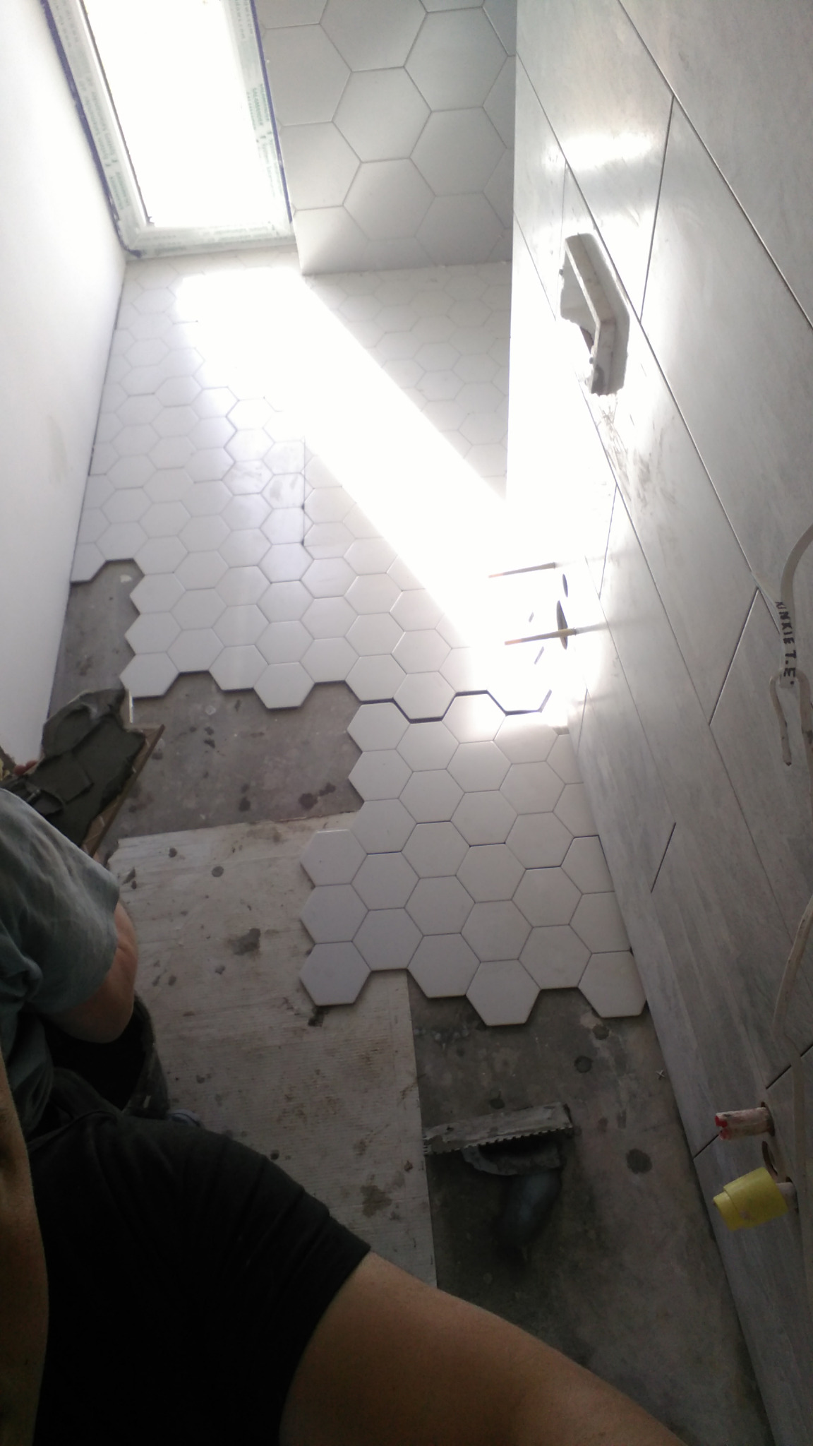 Układanie białych, sześciokątnych płytek podłogowych w łazience, widoczne narzędzia glazurnicze i fragment instalacji wodnej, w tle jasne płytki ścienne.