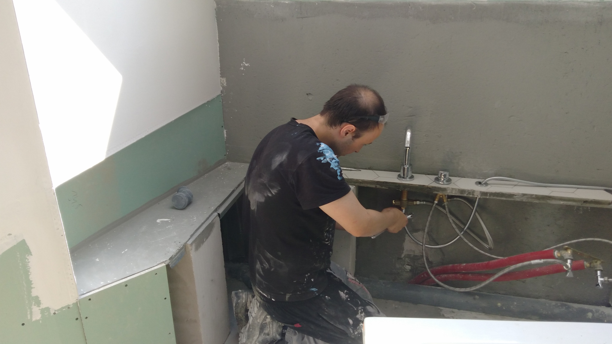 Hydraulik w trakcie montażu baterii umywalkowej w surowym wnętrzu łazienki, widoczne rury doprowadzające wodę i płyty gipsowo-kartonowe na ścianach.