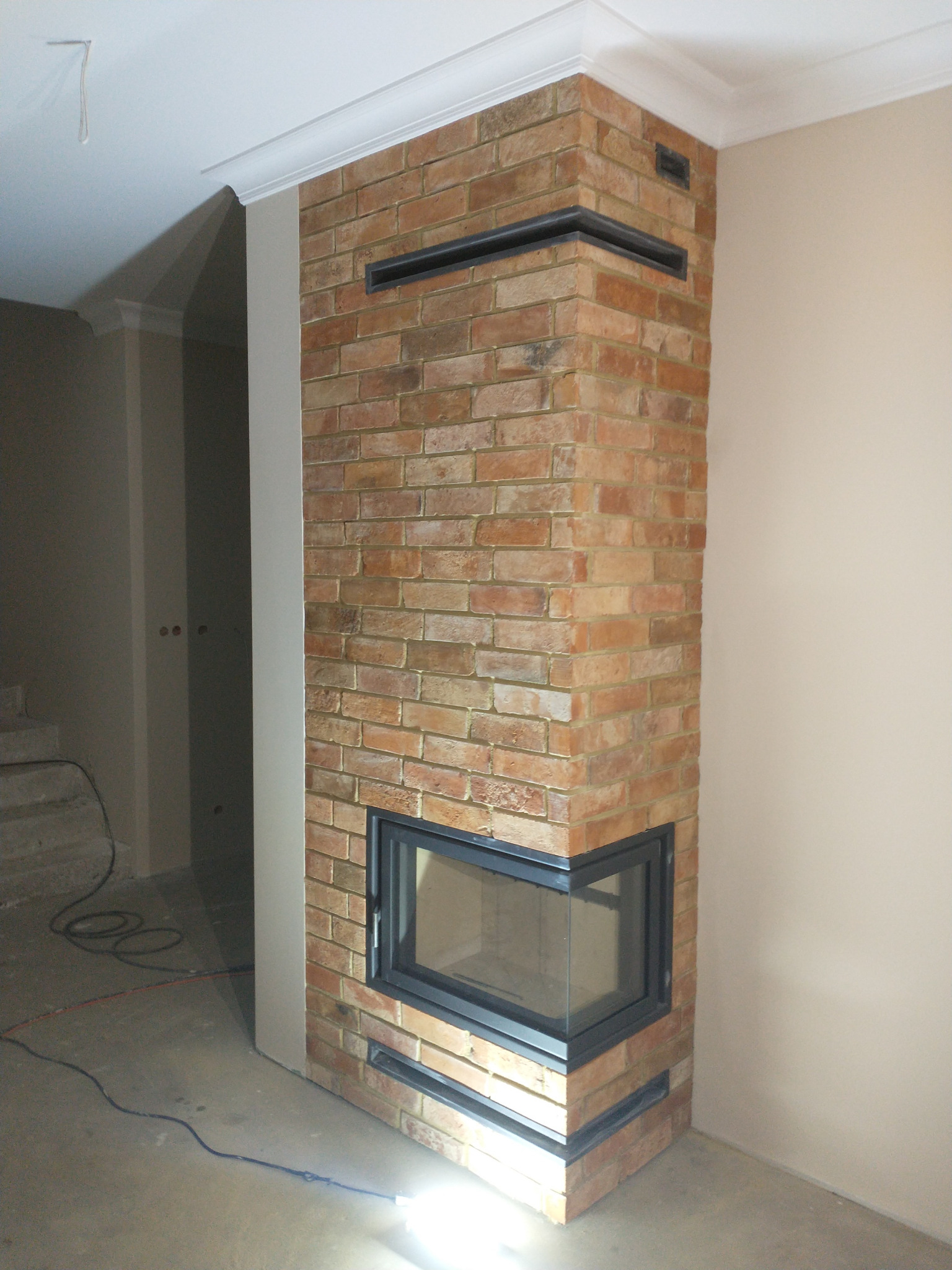 Murowany kominek narożny z cegły klinkierowej, z czarną obudową w surowym wnętrzu, widoczne przewody elektryczne na podłodze.