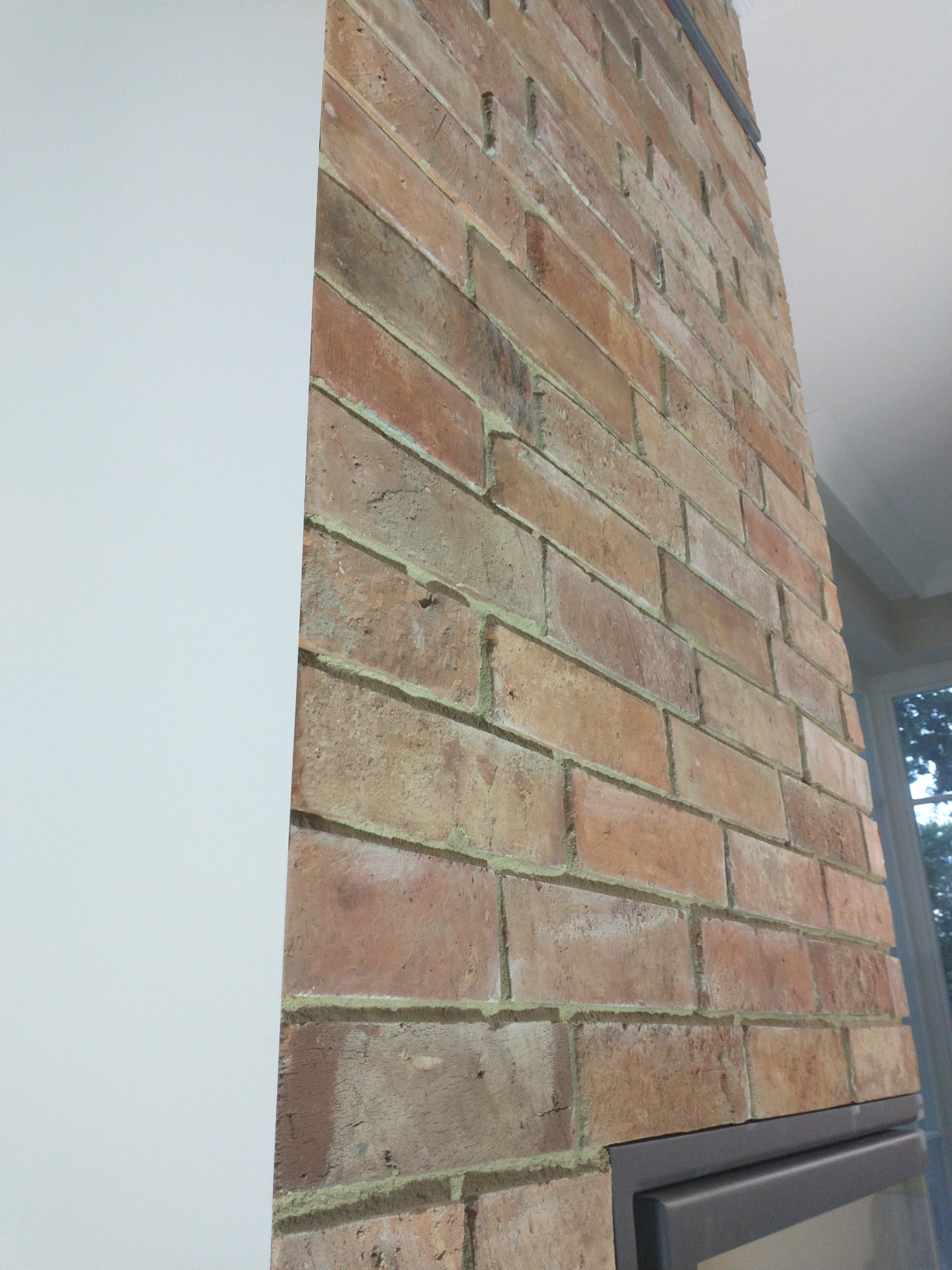 Fragment ściany z cegły klinkierowej wewnątrz pomieszczenia, widoczny styk z białą ścianą i fragment okna.