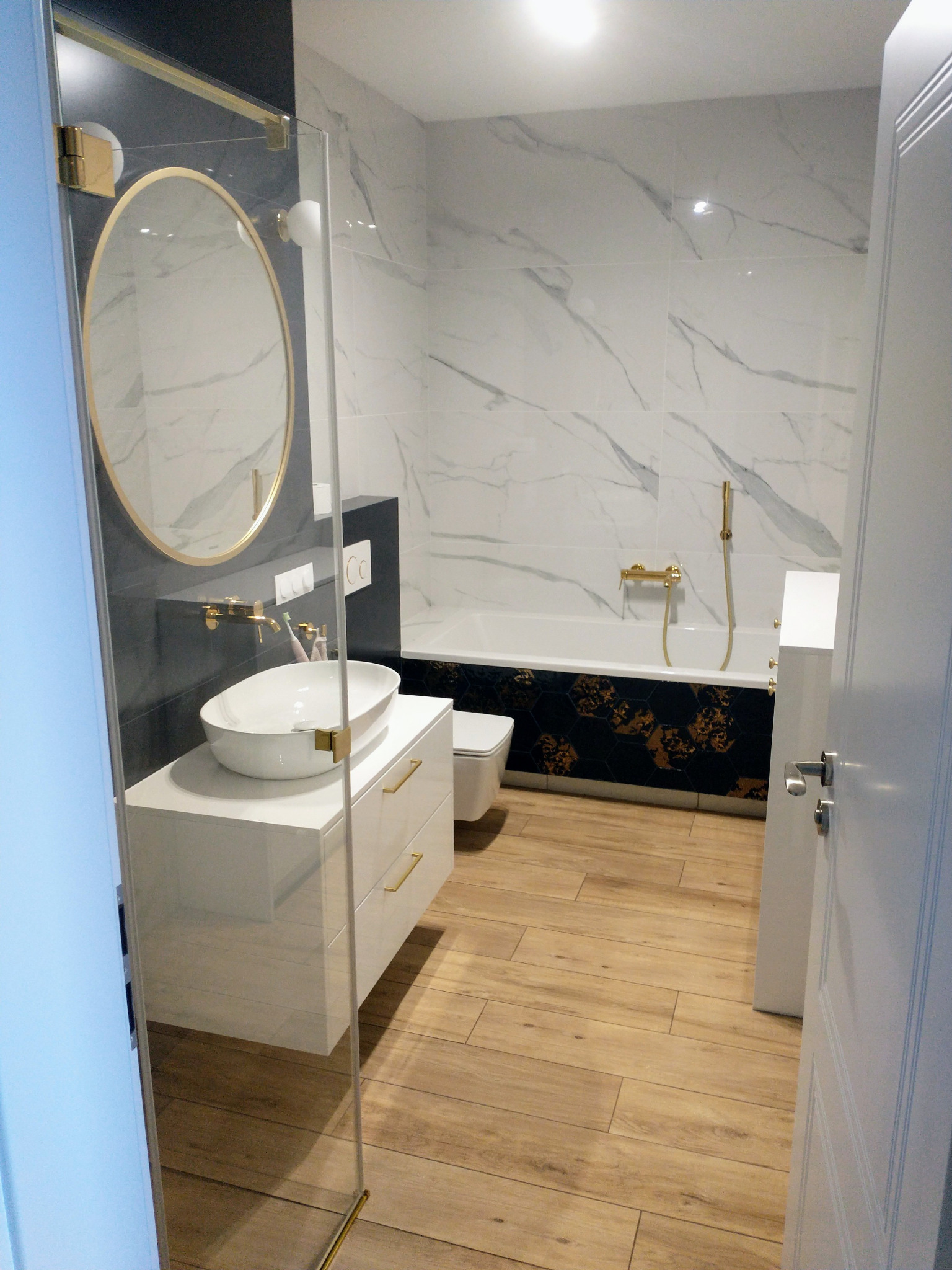 Nowoczesna łazienka z wanną obudowaną czarno-złotymi płytkami, umywalką nablatową na białej szafce z złotymi uchwytami i okrągłym lustrem w złotej ramie, kabina prysznicowa ze złotymi okuciami...