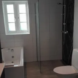 Nowoczesna łazienka z białymi płytkami, kabiną prysznicową typu walk-in, wanną z hydromasażem i toaletą, wykończona czarnym marmurem.