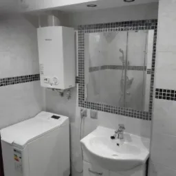 Wykończona łazienka w odcieniach szarości i bieli z pralką, umywalką z szafką, lustrem w mozaikowej ramie i piecem gazowym.