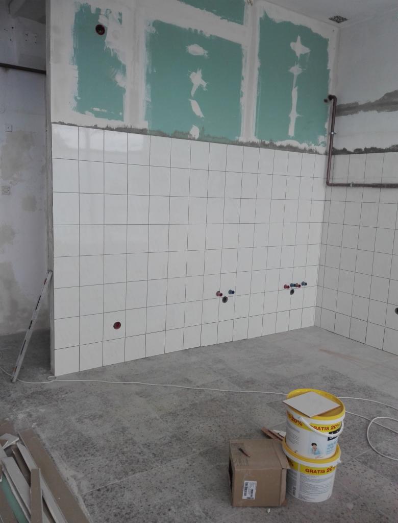 Wnętrze w trakcie remontu: białe, kwadratowe płytki ułożone na ścianie do połowy wysokości, widoczne wyprowadzenia instalacji, ściany powyżej przygotowane pod dalsze prace, na podłodze dwa wiadra...