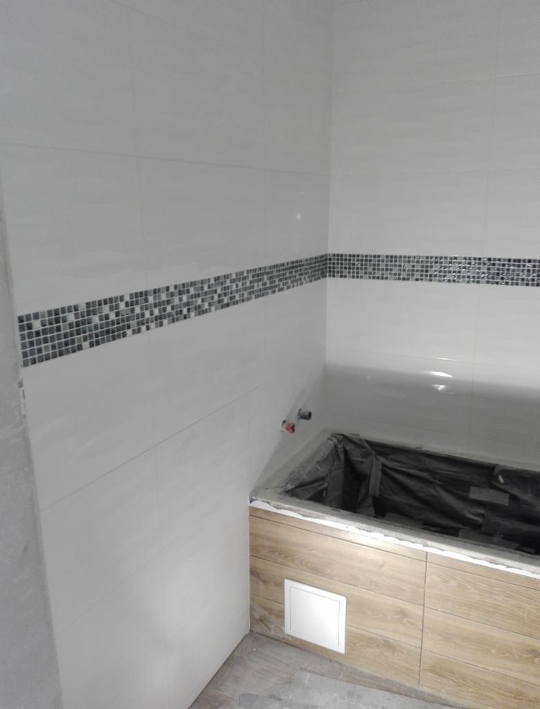Wnętrze łazienki w trakcie prac wykończeniowych: białe płytki ścienne z dekoracyjnym pasem mozaiki, obudowa wanny z paneli imitujących drewno, widoczne rury instalacji wodnej.