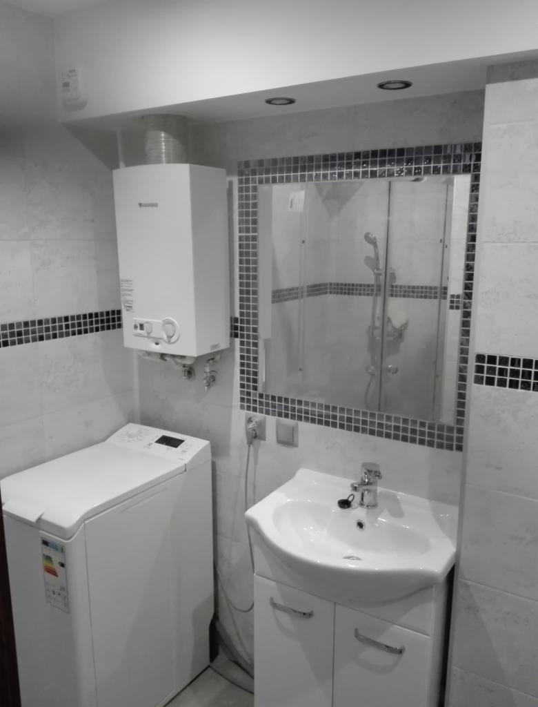 Wykończona łazienka w odcieniach szarości i bieli z pralką, umywalką z szafką, lustrem w mozaikowej ramie i piecem gazowym.