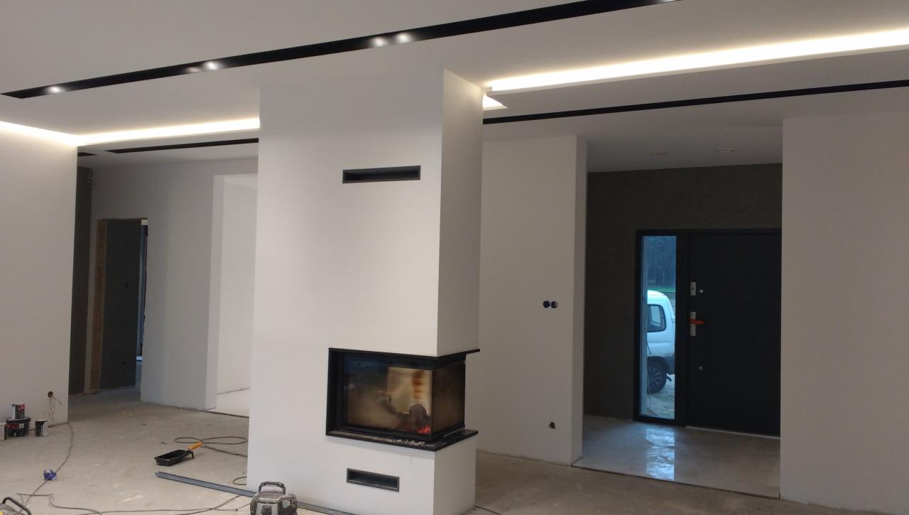 Wnętrze w trakcie wykańczania: białe ściany, czarne listwy sufitowe z oświetleniem LED, nowoczesny kominek narożny z paleniskiem, widoczne narzędzia malarskie na podłodze, drzwi wejściowe...