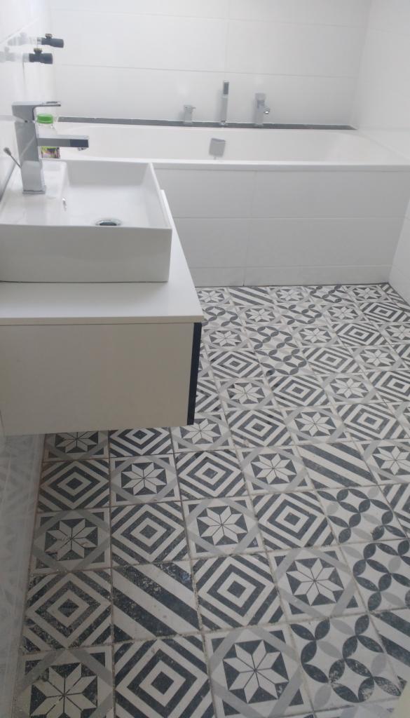 Biała łazienka z umywalką nablatową i wanną w zabudowie, wzorzyste płytki podłogowe w odcieniach szarości i bieli.