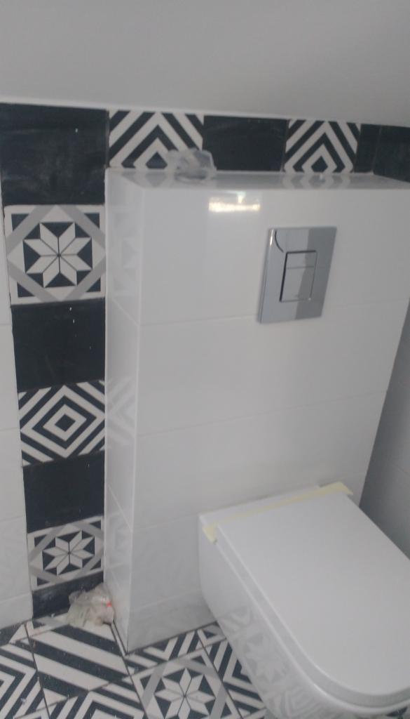 Nowoczesna toaleta podwieszana z białym stelażem ukrytym za płytkami, z chromowanym przyciskiem spłukującym i dekoracyjnymi płytkami podłogowymi oraz ściennymi w czarno-białe wzory geometryczne.