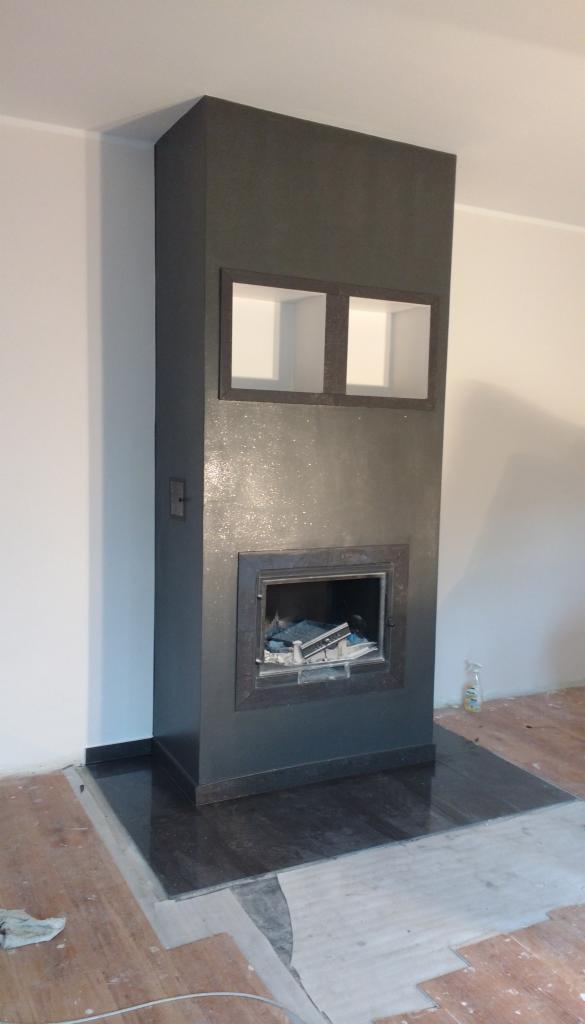 Obudowa kominka w kolorze grafitowym z dwoma małymi okienkami u góry, osadzona na czarnym marmurze, w trakcie prac wykończeniowych podłogi.