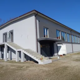 Dwukondygnacyjny budynek z elewacją w odcieniach szarości, z zewnętrznymi betonowymi schodami prowadzącymi do otwartych drzwi na piętrze, widoczne ślady prac budowlanych na gruncie.