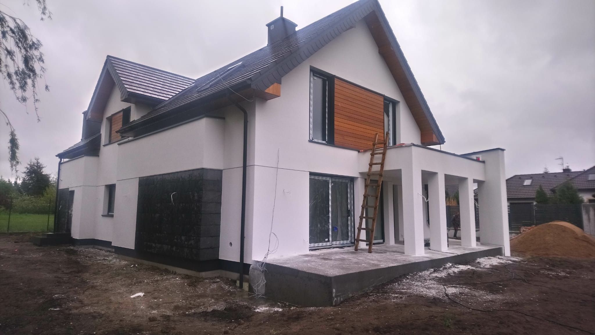 Dwukondygnacyjny dom z białym tynkiem i elementami z ciemnego kamienia, drewnianą fasadą i drewnianą drabiną opartą o ścianę, ukazujący etap prac wykończeniowych na zewnątrz budynku w pochmurny dzień.