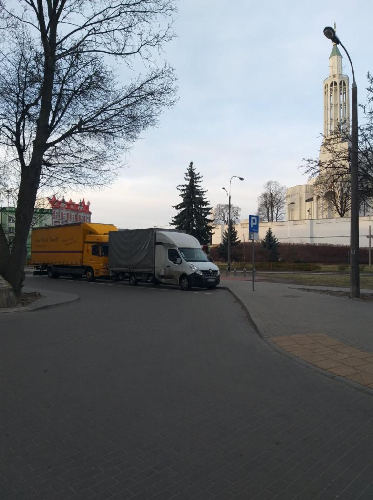 Dwa zaparkowane pojazdy dostawcze: żółta ciężarówka i biały bus z plandeką na tle miejskiego krajobrazu z kościołem w oddali.