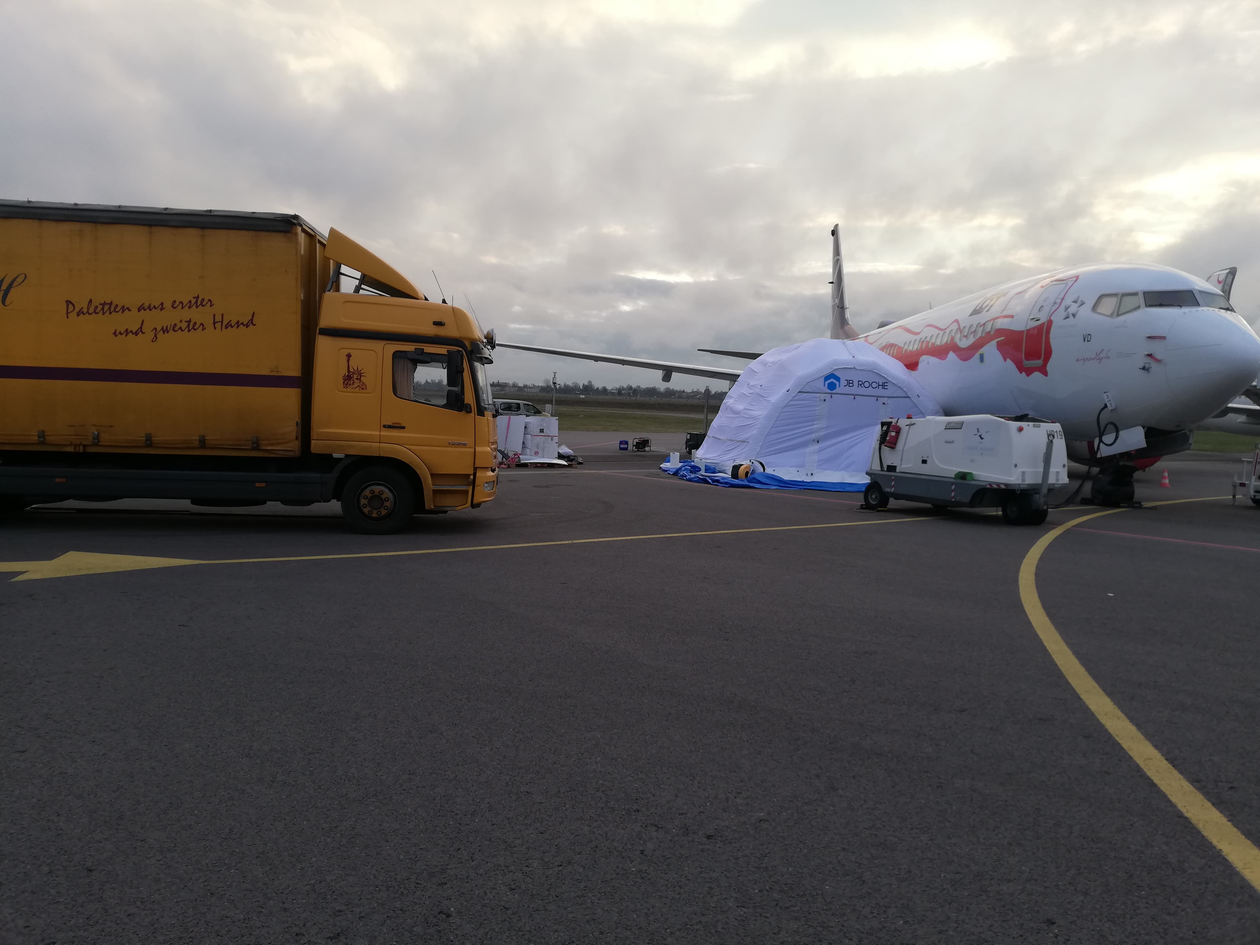 Żółta ciężarówka z napisem obok samolotu i namiotu na płycie lotniska. Transport specjalny, nietypowy ładunek.