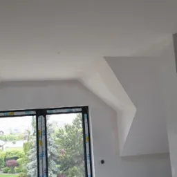 Poddasze z oknem dachowym po nałożeniu pierwszej warstwy gładzi, widoczne niedoskonałości i nierówności na ścianach, przygotowane do dalszych prac wykończeniowych.