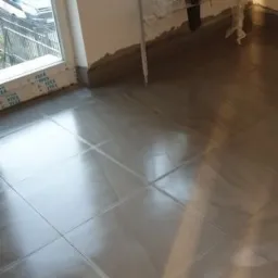 Świeżo położone, błyszczące, szare płytki podłogowe w pokoju z dużym oknem, widoczny fragment ściany z niedokończonym wykończeniem przy podłodze oraz suszarka na pranie zabezpieczona folią.