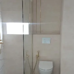 Nowoczesna łazienka z kabiną prysznicową typu walk-in, złotymi akcentami armatury i drewnopodobną podłogą, widok na toaletę podwieszaną i przyciski spłukujące.