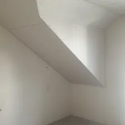 Niedokończone poddasze z zamontowanymi płytami gipsowo-kartonowymi na skośnym suficie i ścianach, widoczne profile i resztki materiałów na podłodze.